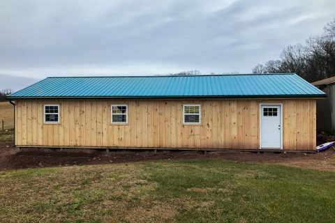 Back of horse barn with 3 windows, a white door, light-colored wood siding, and teal roofing.