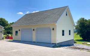 2 car concrete foundation garage with 2 white garage doors, light yellow vinyl siding, light shingle roofing, and 3 exterior lights.