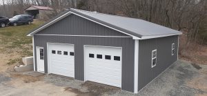 2 car pole barn garage with dark gray metal siding, white trim, 2 white garage doors, a white single entry door, windows with white trim, and gray metal roofing.