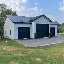 3 car detached garage with white siding, black metal roofing, and black garage doors. The garage door in the middle is larger than the 2 on both sides.