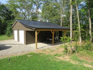 Front and left side view of a 2 car garage with 2 white garage doors, brown metal siding, black metal roofing, a white single entry door, and an attached car port cover.