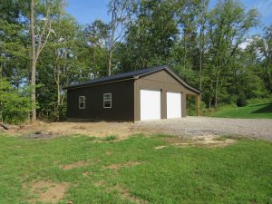 Back and side view of a 2 car garage with 2 white garage doors, brown metal siding, black metal roofing, and windows with white trim.