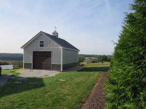 Front view of a two tone garage with light gray and dark gray metal siding, a brown garage door, white trim, dark gray metal roofing, and a cupola.