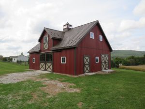 Front and right side view of a 2-story horse barn with red metal siding, brown metal roofing, 2 sets of Dutch barn doors, a single entry Dutch barn door, and a cupola.