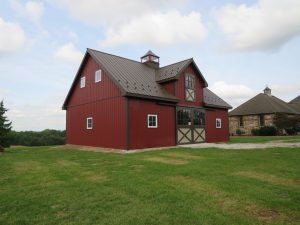 Front and leftside view of a 2-story horse barn with red metal siding, brown metal roofing, windows with white trim, a set of Dutch barn doors, and a cupola.