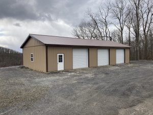 3 car pole barn garage with brown metal siding, 3 white garage doors, a white single entry door with windows, dark brown trim, and dark brown metal roofing.