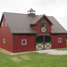 Large barn with red siding, brown metal roofing, a cupola, windows with white trim, and a set of double doors with windows.